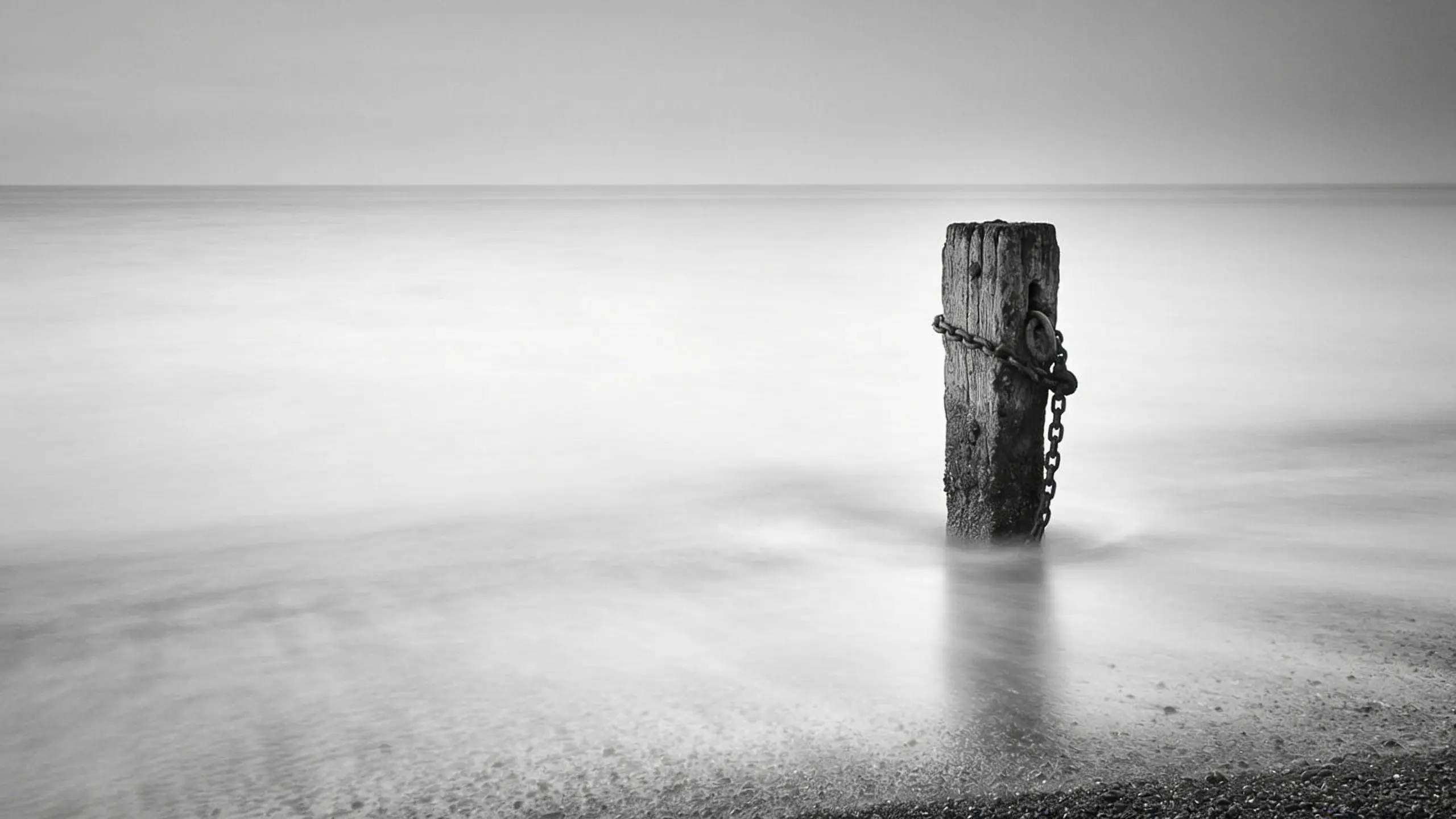 Black and white long exposure: a single weathered wooden post in shallow water, the horizon dissolving into haze
